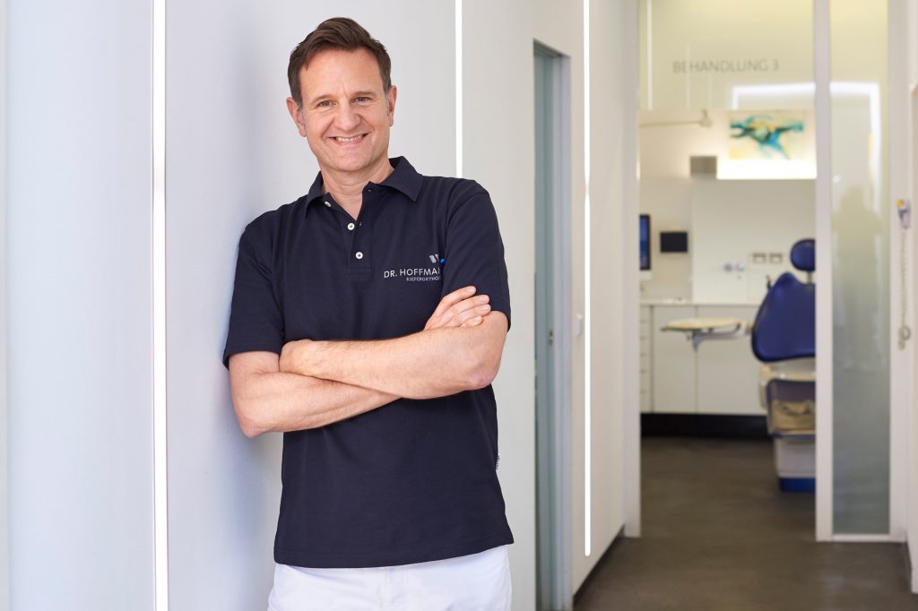 Kieferorthopäde Dr. Hoffmann lehnt an der Wand und schaut lachend in Kamera